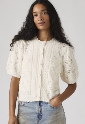 Γυναίκα με μακριά, σγουρά μαλλιά που φοράει λευκό πλεκτό ζακέτα με κουμπιά και φουσκωτά μανίκια, καθώς και ανοιχτό μπλε τζιν, με το χέρι στην τσέπη.