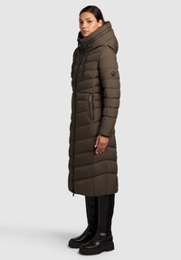 Garšs, polsterēts, tumšzaļš mētelis ar augstu apkakli un kapuci, ar rāvējslēdzēju un sānu kabatām, apvienots ar melniem ceļgalu augstiem zābakiem.