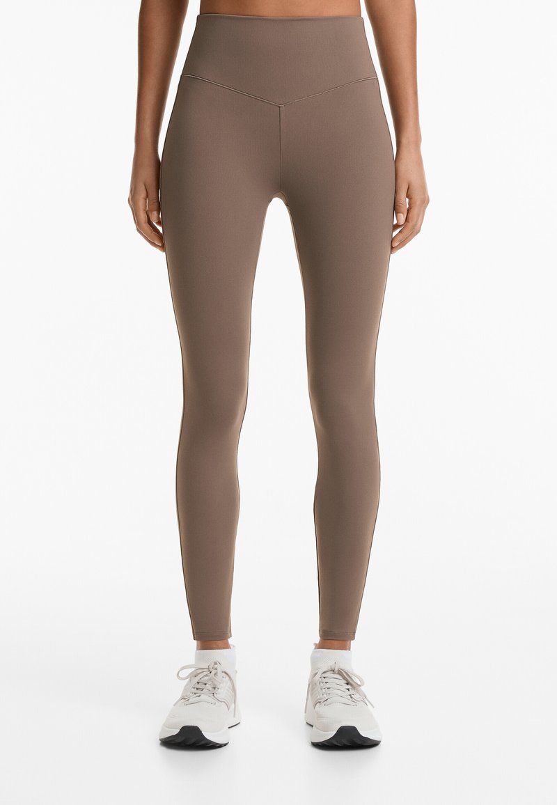 Braune hochtaillierte Leggings aus dehnbarem Stoff, mit glatter Textur und figurbetonender Silhouette, kombiniert mit weißen Sneakern.