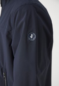 Gros plan sur la manche d'une veste bleu foncé avec un patch logo rond "Save The Duck" représentant une icône de canard.