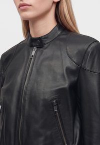 Femme portant une veste en cuir noire avec une fermeture éclair devant et un col à boutons pression, visible des épaules jusqu'au milieu de la poitrine.