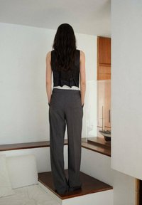 Schwarze ärmellose Weste mit einer Schleife auf dem Rücken, getragen über einem weißen Oberteil. Graue hoch taillierte Hose mit seitlichen Taschen, stehend auf einer Holzoberfläche.