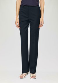 Pantalon à motif bleu marine avec une coupe ajustée, jambes droites et texture subtile ; porté avec des sandales à talons blancs à lanières.