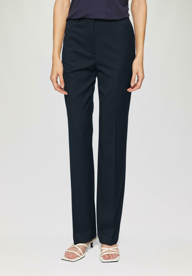 Pantalon à motif bleu marine avec une coupe ajustée, jambes droites et texture subtile ; porté avec des sandales à talons blancs à lanières.