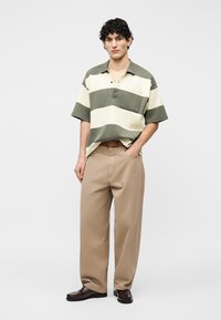 Νέος άνδρας που στέκεται, φοράει πράσινο και κρεμ ριγέ πόλο πουκάμισο, μπεζ φαρδιά παντελόνια, καφέ ζώνη και σκοτεινά καφέ παπούτσια.