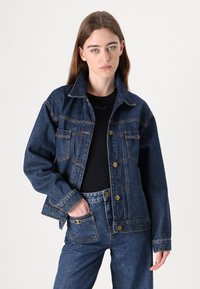 Mörkblå denimjacka med spetsig krage, två bröstfickor och guldfärgade detaljer. Avslappnad passform med sömmaningar och manschetter.