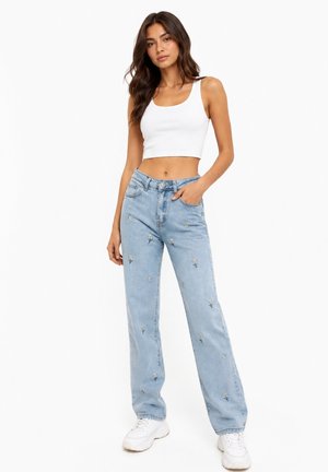 Kvinde iført hvid cropped tanktop og lyseblå jeans med lille hvid blomst broderi, stående med den ene hånd i lommen.