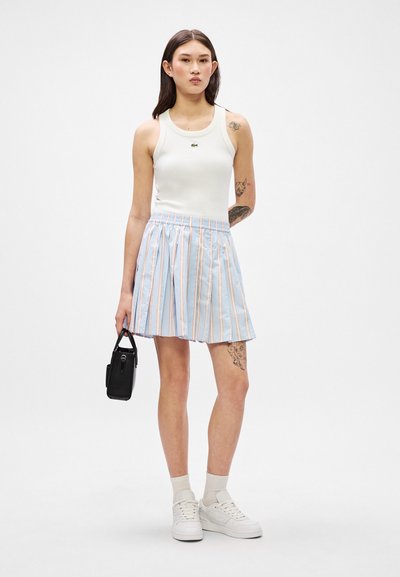 Débardeur blanc avec un petit logo, jupe plissée bleu clair avec des rayures roses et blanches, sac à main noir, et baskets blanches avec des chaussettes à côtes.
