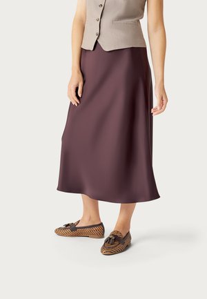 Donna che indossa un gilet beige senza maniche con bottoni, una gonna midi viola e mocassini marroni con motivo e nappine, in piedi su uno sfondo chiaro.