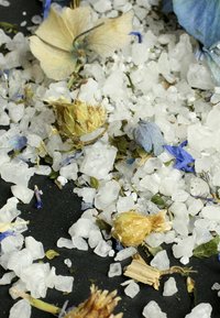 Fiori e erbe essiccate mescolati con cristalli di sale bianco grossolano su uno sfondo nero. I colori principali includono blu, giallo e verde.