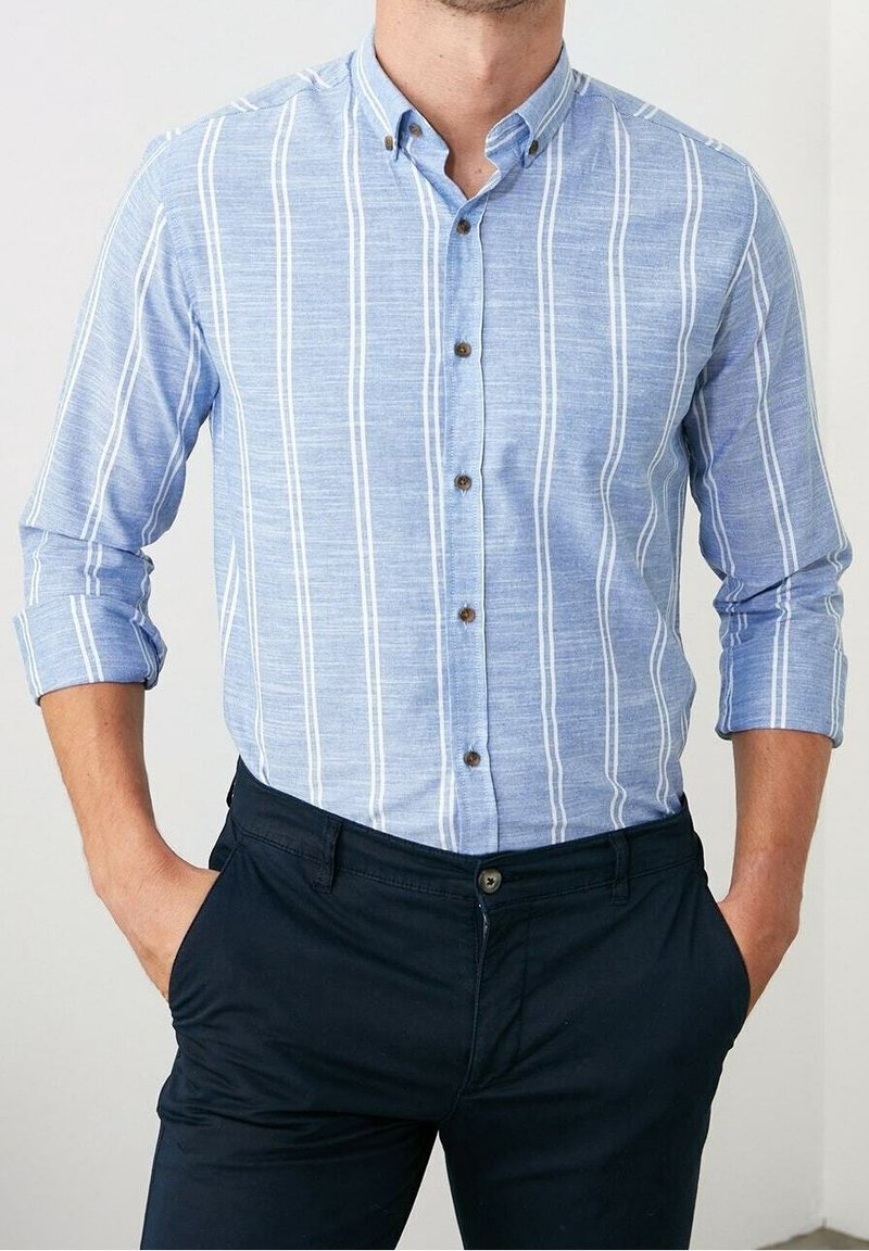 Homme portant une chemise bleu clair à rayures verticales blanches et un pantalon bleu foncé, mains dans les poches, manches retroussées.