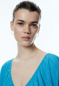 Blauwe geplooide top met een diepe V-hals en korte mouwen. Gladde textuur, geaccentueerd met kleine oorringen. Witte achtergrond.