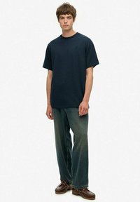 Jonge man staand, gekleed in een effen donkerblauwe T-shirt, losse vervaagde blauwe spijkerbroek en bruin leren schoenen tegen een witte achtergrond.