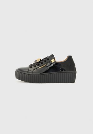 Zwarte platform sneaker met dikke geribbelde zool, glanzende leren details, zwarte veters en een gouden ritssluiting en bij-vormige bedel aan de zijkant.