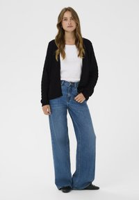 Čierny pletený cardigan cez bielu košeľu, doplnený o modré nohavice s vysokým pásom a širokými nohavicami. Model stojí vzpriamene na jednotnom pozadí.