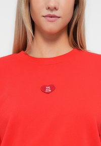 Sweatshirt en coton rouge avec un patch brodé en forme de cœur affichant le texte "THE V.I.P. CLUB" en blanc. Design simple à col ras du cou.