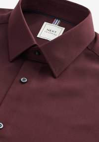 Bordeauxfarbendes Hemd aus strukturiertem Stoff, mit Button-Down-Kragen und grauen Knöpfen. Das Etikett zeigt mehrfarbige Streifen und die Marke "NEXT TAILORING".
