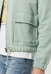 Light green zip-up jacket with flap pocket, elastic cuffs and hem, worn over a white shirt and blue jeans on a person's torso and arm.