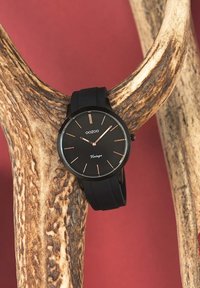 Montre Oozoo noire avec un cadran rond minimaliste, des accents en or rose, un bracelet en silicone, et placée sur un bois de cerf rustique sur un fond rouge.