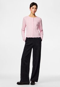 Femeie cu păr scurt și închis la culoare poartă un cardigan roz deschis cu nasturi, pantaloni negri cu picioare largi și pantofi negri, stând cu mâinile în buzunare.