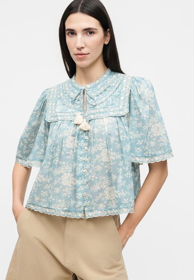 Γυναίκα που φοράει απαλή γαλάζια φλοράλ μπλούζα με κυματιστά τελειώματα, κοντά φουσκωτά μανίκια, κουμπιά μπροστά και κορδόνια με φουντάκια στο γιακά.