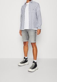 Koszula na guziki w niebiesko-białe paski, zestawiona z jasnoszarymi, podwiniętymi szortami oraz czarnymi trampkami za kostkę z białymi podeszwami.