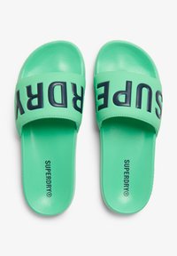 Groene sliders met een gestructureerde afwerking, voorzien van een brede band met "SUPERDRY" in donkere letters over de voorkant. Glad zool.