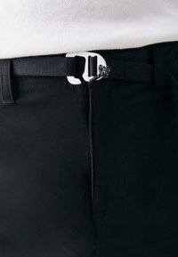 Pantalons noirs avec un tissu texturé, dotés d'une ceinture en sangle noire et d'une boucle en plastique blanche au design unique. Vue en gros plan.