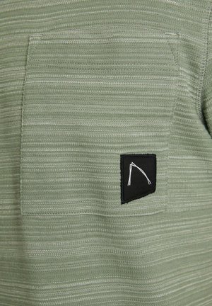 Tejido verde texturizado con líneas horizontales, que presenta un bolsillo negro con un diseño gráfico blanco.