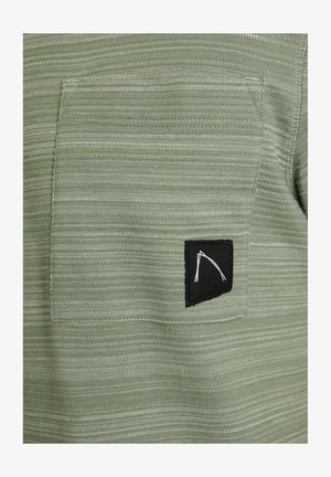 Textúrovaná zelená látka s horizontálnymi pruhmi, s čiernou náprsníkovou vreckom a bielym grafickým dizajnom.