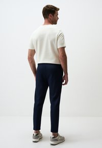 Homme de dos portant une chemise blanche à manches courtes, un pantalon bleu foncé et des baskets de couleur claire sur un fond blanc.