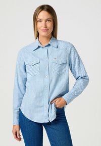 Jasnoniebieska koszula w krate z dwiema kieszeniami na piersi, kołnierzykiem zapinanym na guziki, długimi rękawami i srebrnymi guzikami. Materiał wydaje się miękki i gładki.