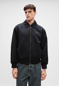 Sort bomberjakke med tekstureret overflade, med lynlås foran, stor krave og ribbede manchetter, båret over blå denimjeans.