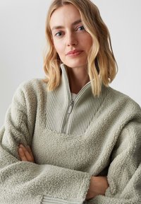 Pullover verde chiaro in pelliccia sintetica con collo alto a coste e chiusura con zip, caratterizzato da una texture morbida e una vestibilità rilassata.