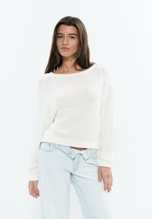 Joven con cabello largo y castaño, vestida con un suéter blanco de punto y jeans azul claro, posando frente a un fondo blanco liso.