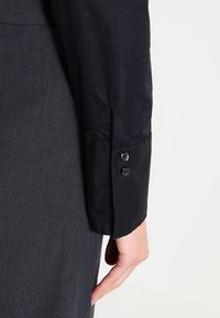 Czarna rękaw koszuli z gładkiego materiału, z dopasowanym mankietem z dwoma guzikami, w połączeniu z ciemnoszarym ubraniem.