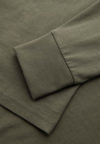 Primer plano de camisa de manga larga verde oliva mostrando un puño y dobladillo doblados con costuras visibles en tela suave.