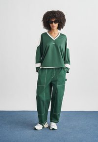 Žena s kučeravými vlasmi, ktorá nosí čierne slnečné okuliare, zeleno-biele športové oblečenie a biele tenisky, stojaca na modrom koberci pred bielou stenou.