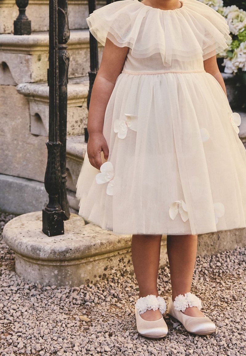 Jeune enfant portant une robe en tulle blanc avec des appliques florales et des chaussures à volant, debout sur du gravier près de marches en pierre et d'une rampe en fer noir.