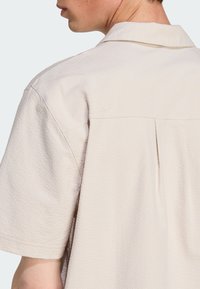 Krótki rękaw, beżowa koszula o teksturowanym wykończeniu, z kołnierzykiem i plisą z tyłu dla dodatkowego detalu.