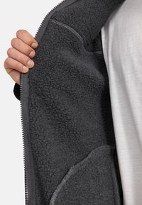 Interior de chaqueta de forro polar gris oscuro con una superficie texturizada, cremallera visible y un bolsillo lateral. Una mano agarra el borde, revelando su suave forro.