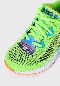 Bright green Skechers Elite Sport sneaker with orange trim, black mesh pattern, and a blue tag reading "Durable rubber bottom".
