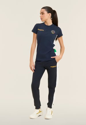 Donna in piedi con la mano in tasca, indossa una t-shirt blu navy del marchio Freddy e pantaloni abbinati con strisce laterali bianche e verdi, oltre a scarpe da ginnastica bianche.