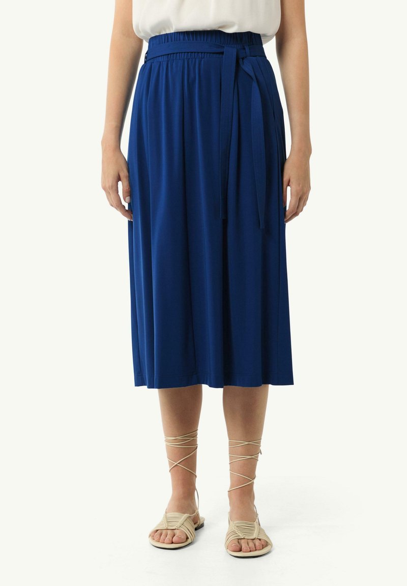 Blauer Midirock mit einem elastischen Taillenbund und passendem Bindeband, aus einem glatten Stoff gefertigt. Kombiniert mit beigen Riemchensandalen.