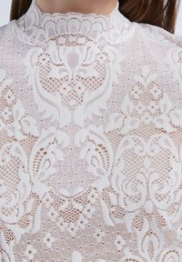 Blouse en dentelle blanche avec des motifs floraux complexes, un col montant, et une surface texturée avec des ouvertures en maille variées.