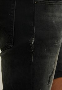 Černé džíny z denimové tkaniny s úzkým střihem, distressed texturou, roztrhanými detaily a vyšitým logem na boku.