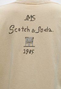 Μπεζ ύφασμα με μαύρο κεντημένο κείμενο "A/S Scotch & Soda" πάνω από ένα μπλε και γκρι κεντημένο έμβλημα και το έτος "1985" από κάτω.