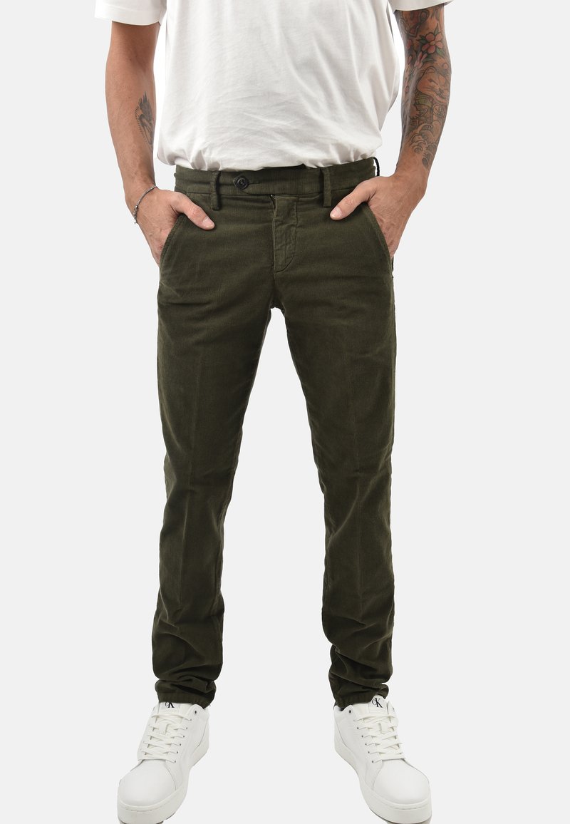 Pantaloni in velluto a coste verde oliva con vestibilità slim, cinque tasche e chiusura con bottone, abbinati a sneakers bianche e una t-shirt bianca semplice.