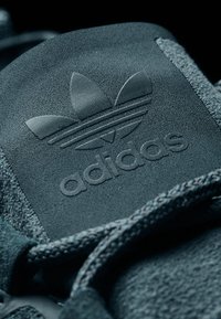 Tmavě šedý semišový jazyk tenisky s embosovaným logem tří pruhů a textem "adidas", s plochou tkanou šňůrkou a texturovaným povrchem.