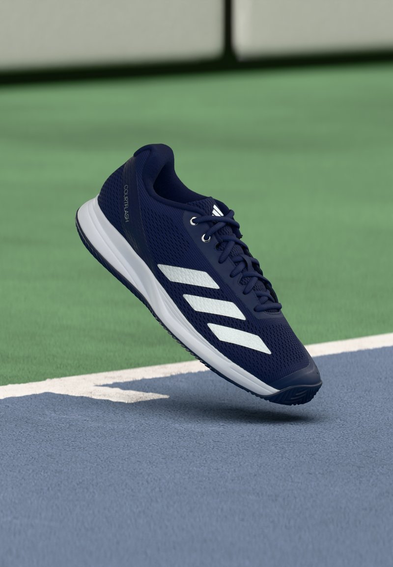 Chaussure de tennis Adidas CourtFlash bleu marine avec des rayures blanches, en suspension au-dessus d'une surface de court de tennis bleue et verte.